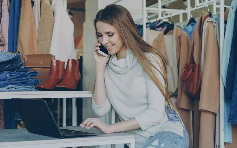 Woman using a laptop while talking on the phone in a clothing shop.