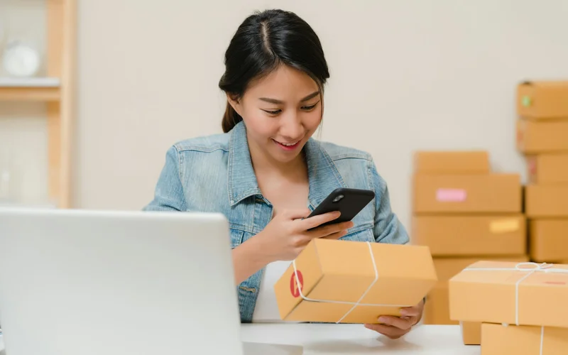 Woman checking her phone beside packages and a laptop.